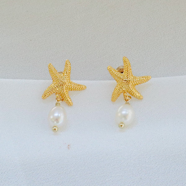 Pearlpals Gold-plated earrings with starfish-shaped tops and a pearl drop, displayed on a soft white fabric background.