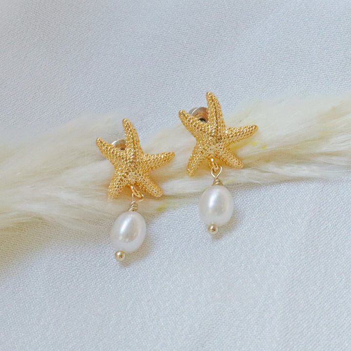 Pearlpals Gold-plated earrings with starfish-shaped tops and a pearl drop, displayed on a soft white fabric background.