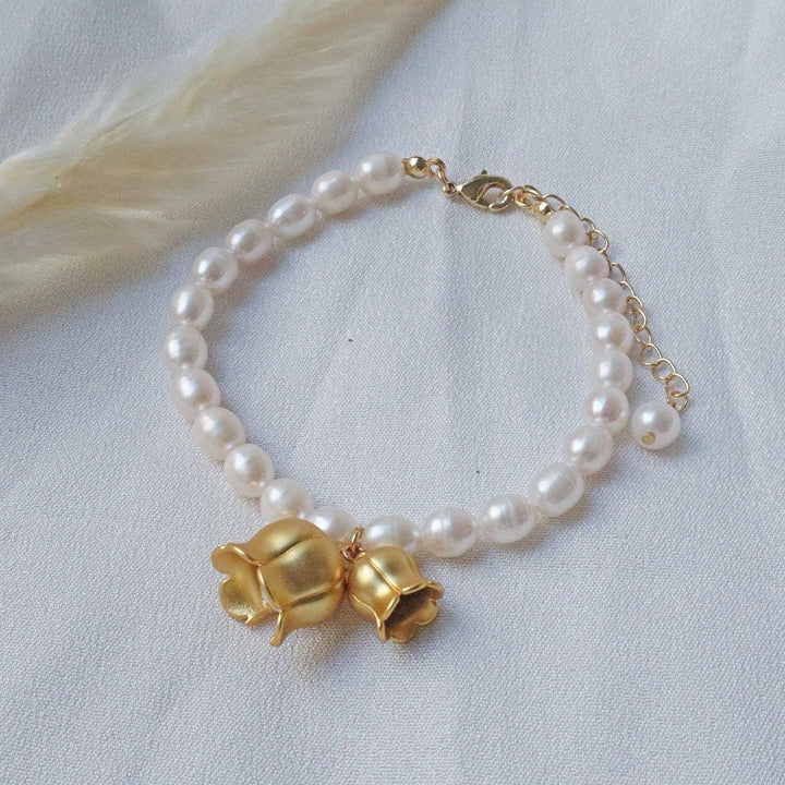 Pearlpals A gold-plated bracelet with pearls, featuring two gold lily of the valley charms each encasing a pearl, displayed on a soft white fabric background.