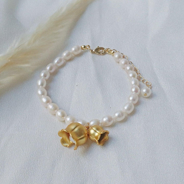 Pearlpals A gold-plated bracelet with pearls, featuring two gold lily of the valley charms each encasing a pearl, displayed on a soft white fabric background.