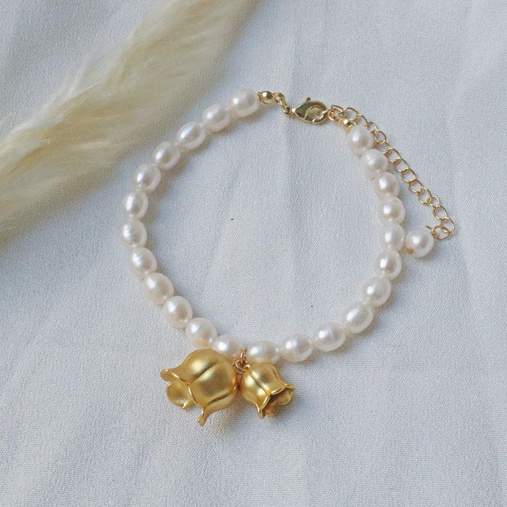 Pearlpals A gold-plated bracelet with pearls, featuring two gold lily of the valley charms each encasing a pearl, displayed on a soft white fabric background.