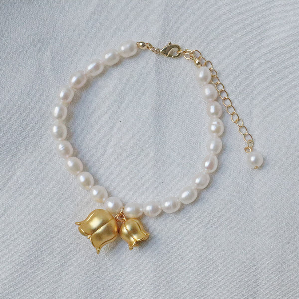 Pearlpals A gold-plated bracelet with pearls, featuring two gold lily of the valley charms each encasing a pearl, displayed on a soft white fabric background.